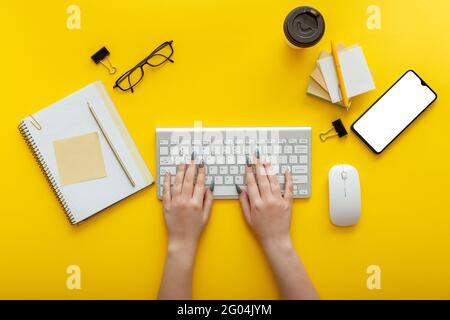 Woman typing on computer keyboard at workplace on color yellow background. Office desktop workspace with female hands, cup of coffee smartphone mockup Stock Photo