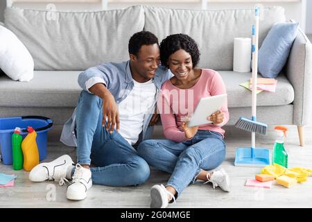 Smiling black couple with cleaning tools resting, using digital tablet Stock Photo