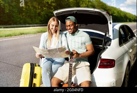 Multiracial couple going on road trip in car, looking at map, taking break from driving, sitting in auto trunk together Stock Photo