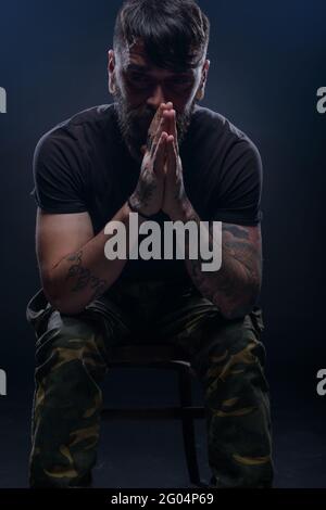 Closeup dark picture of a bearded male model in black t-shirt  sitting on a chair and praying with his tattooed hands Stock Photo