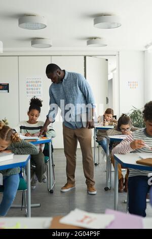 Vertical full length portrait of male African-American teacher helping kids in school classroom Stock Photo