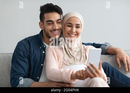 Cheerful Pregnant Middle Eastern Spouses Browsing Internet On Smartphone Together At Home Stock Photo