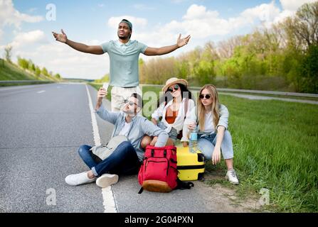 Unhappy millennial tourists sitting on roadside, cannot catch car, waiting for too long, having no smartphone connection Stock Photo