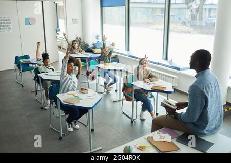 Diverse group of children raising hands in school classroom while sitting at desks, copy space Stock Photo