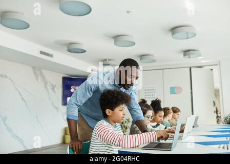 Wide angle side view portrait of young boy using computer in IT class with male teacher helping him, copy space
