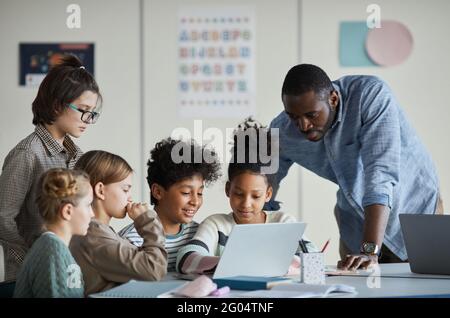 Diverse group of children with male teacher using laptop together in modern school classroom Stock Photo