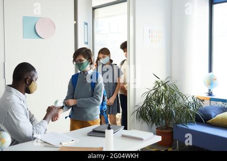 Group of children wearing masks entering classroom in school with teacher checking temperature with infrared thermometer, covid safety Stock Photo