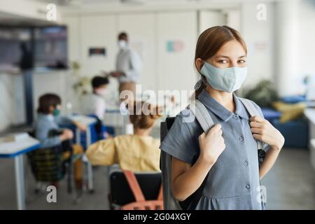 Waist up portrait of cute girl wearing mask in school classroom and looking at camera , covid safety measures, copy space Stock Photo