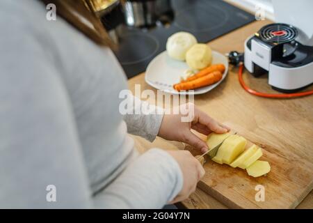 over the shoulder view on hands of unknown woman with knife on board in the kitchen cutting pieces of potato for the vegan or vegetarian soup cooking Stock Photo