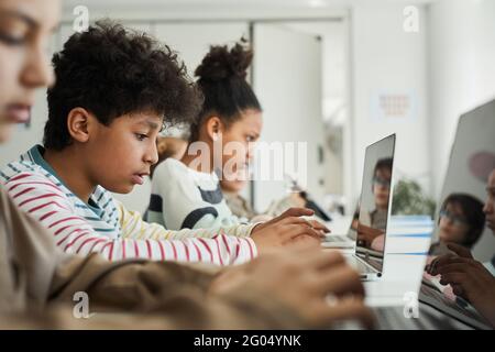 Side view at diverse group of children sitting in row at school classroom and using computers Stock Photo