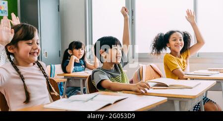 Elementary age Asian student boy raised hands up in class. Diverse group of pre-school pupils in elementary age in education building school. Volunteering and participating classroom concept. Stock Photo