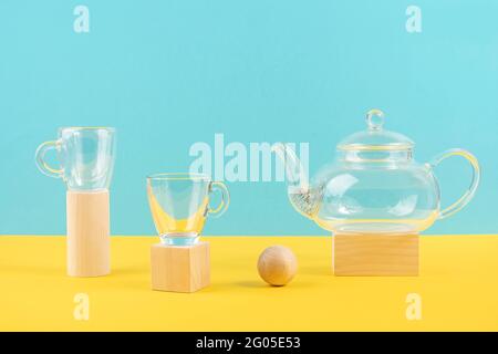 Empty glass transparent tea set. Teapot and and two cups standing on wooden geometric shapes, yellow and blue background. Front view. Stock Photo