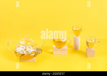 Three cups of camomile tea, transparent teapot on wood geometric shape, yellow background. Creative concept Natural Chamomile Tea. Top view Flat lay. Stock Photo