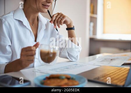 Smiling businesswoman drinking latte and using laptop at work Stock Photo