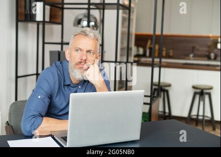 Pensive mature man solving difficult business task sitting at the desk with a laptop and looks away, senior male entrepreneur thinking Stock Photo