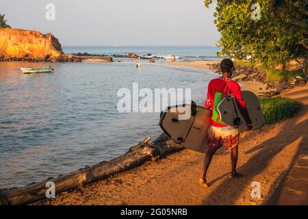 Surfer carries two surfboards along path beside Parrot Rock Island, after sunrise, Mirissa Beach, Southern Province, Sri lanka Stock Photo