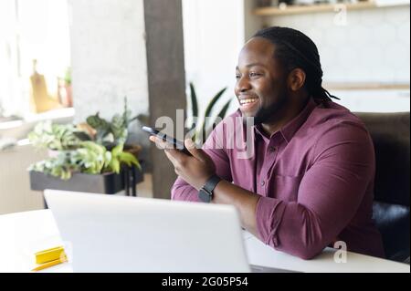 Smiling African-American man with locks hair sending voice message sitting at home office, positive black guy in casual shirt using voice recognition app, recording audio report, side view Stock Photo
