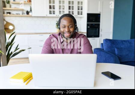 Guy in wirelessheadset using laptop sitting in the kitchen home. Remote work concept Stock Photo