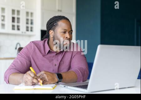 Serious and focused African-American guy watching webinars on the laptop and taking notes, studying remotely from home, writing down some important things, working on new project Stock Photo