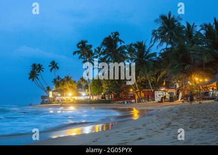Beach scene with palm trees and shacks after dark lit by yellow street lights, Hikkaduwa, Southern province, Sri Lanka Stock Photo