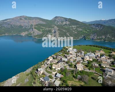 AERIAL VIEW. Village perched 250m-high above Lake Serre-Poncon. Le Sauze-du-Lac, Hautes Alpes, France. Stock Photo
