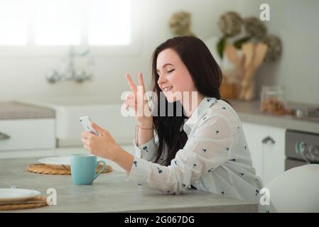 Young brunette woman student is sitting in kitchen with mobile smartphone, smiling and communicating through the screen. Modern gadgets in our life Stock Photo