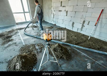 Unknown worker in overalls using pneumatic conveyor and tacheometer in flat of building under construction. Preparing for making cement floor screed Stock Photo