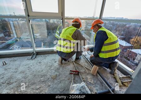 Two workers in protective clothing and safety helmets installing plastic pipes using soldering iron on balcony of flat of building under construction Stock Photo