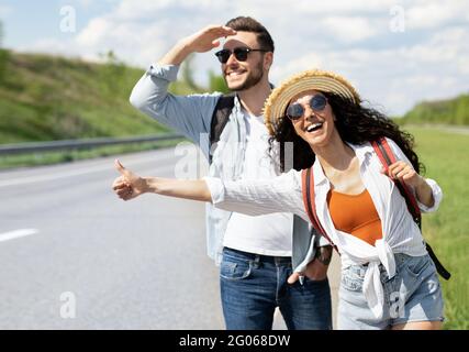Happy young coupe flagging down car, needing ride, hitchhiking on roadside outdoors Stock Photo