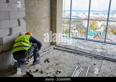 Builder in workwear and safety helmet is installing plastic pipes in flat of building under construction with panoramic windows. High-rise house Stock Photo