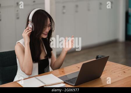 Smiling girl student wear wireless headphone study online with teacher, happy young asian woman learn language listen lecture watch webinar write Stock Photo