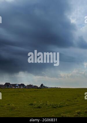A distant inky storm cloud, hurling rain onto a group of houses, advances to obscure a summery sky with sunlit fluffy clouds, over an empty field. Stock Photo