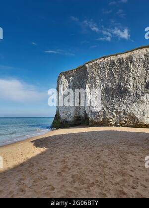 Gentle waves roll in from a blue sea to break on golden sand under a white chalk cliff with a summery blue sky with fluffy white clouds overhead. Stock Photo