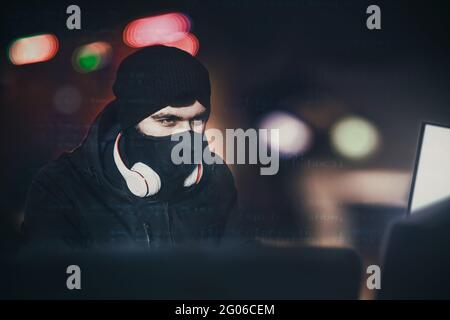 Angry computer hacker weared balaclava stealing data from laptop in front of black background and blue light. close portrait. hack concept Stock Photo