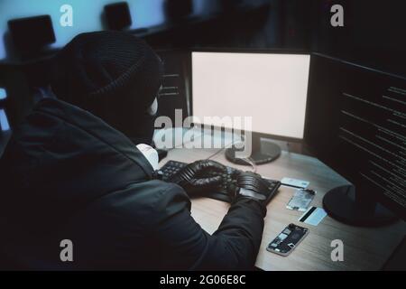 Shot from the Back to Hooded Hacker Breaking into Corporate Data Servers from His Underground Hideout. Place Has Dark Atmosphere, Multiple Displays Stock Photo