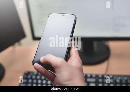 Person holding a smart phone and pointing her finger at the blank screen against a computer keyboard,monitor on wooden table. business and technology Stock Photo
