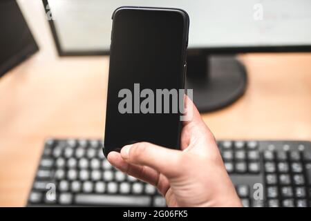 Person holding a smart phone and pointing her finger at the blank screen against a computer keyboard,monitor on wooden table. business and technology Stock Photo