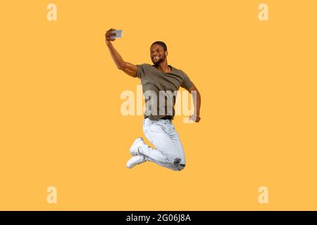 Positive african american man jumping and making selfie on smartphone over yellow background Stock Photo