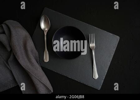 Empty black ceramic bowl, vintage cutlery and a gray linen fabric over a dark slate background Stock Photo