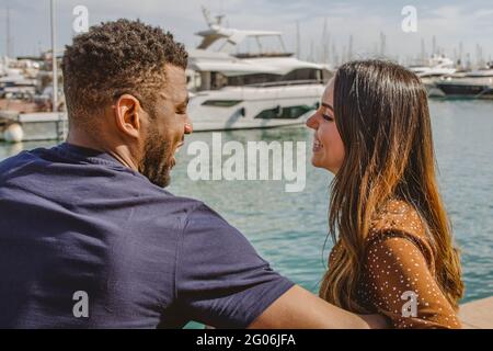 Multiethnic Young Couple Laughing and Looking Each Other with Love Having Fun Sitting at Harbor Stock Photo