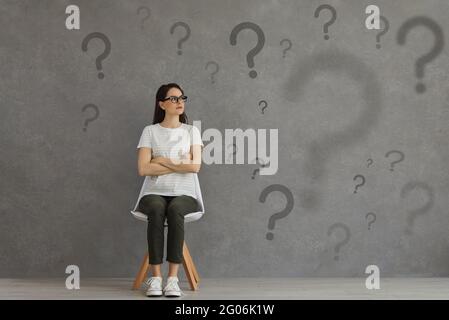 Confused woman sitting on chair and looking at lots of question marks on grey background Stock Photo