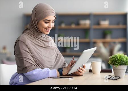 Smiling muslim woman using tablet sitting at desk Stock Photo