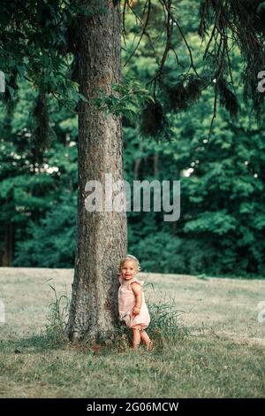 Cute smiling happy baby girl in pink dress standing by tall tree and exploring learning nature world. Small tiny kid child kid toddler in big world Stock Photo