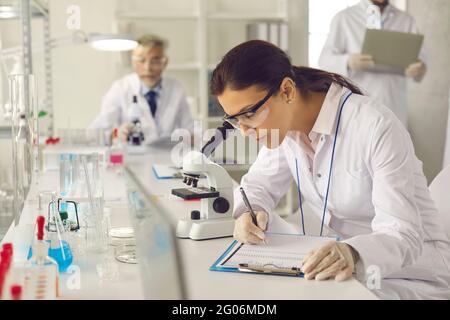 Scientist in lab coat, goggles and gloves looking in microscope and taking notes Stock Photo