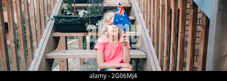 Funny Caucasian girl holding Australian flag. Child sitting on backyard at home and holding Australia flag. Kid citizen celebrating Australia Day holi Stock Photo
