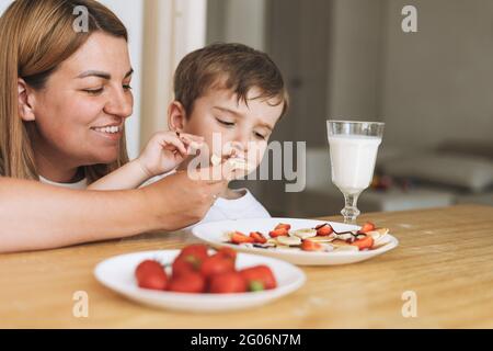 Cute toddler boy with his mother having breakfast with puncakes and berries in bright kitchen at home Stock Photo