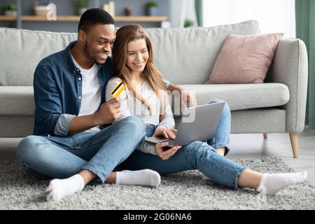 Online Shopping. Cheerful Mixed Couple Using Laptop And Credit Card At Home Stock Photo
