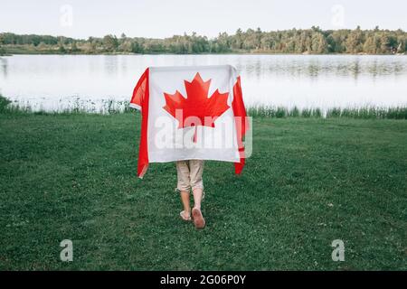 Girl wrapped in large Canadian flag by Muskoka lake in nature. Canada Day celebration outdoor. Kid in large Canadian flag celebrating national Canada Stock Photo