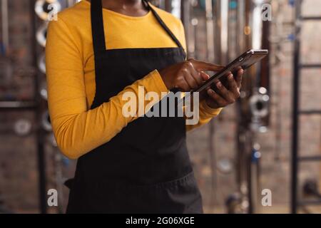 Mid section of african american female worker wearing apron using digital tablet at gin distillery Stock Photo