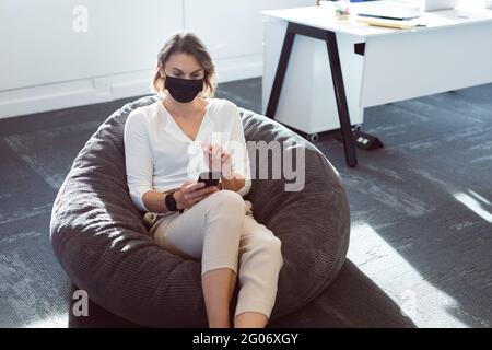 Caucasian businesswoman wearing face mask, sitting in armchair, using smartphone Stock Photo